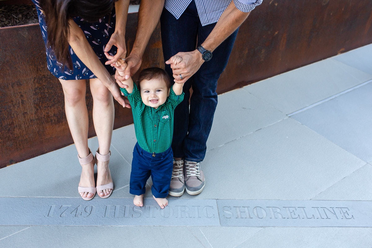 pick a family photographer who can capture candid moments like this image of a mother and father helping baby son stand on sidewalk. Image by best Alexandria, VA baby photographer Julie Kubal
