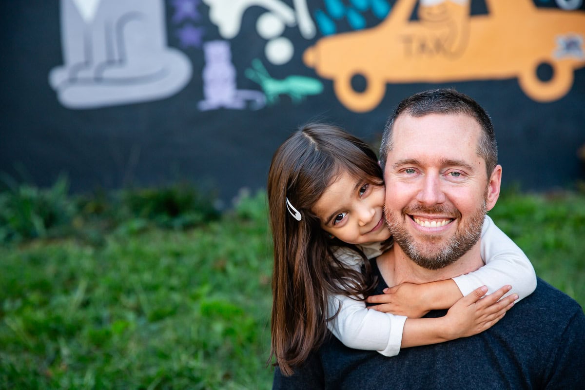 Little girl embraces her father from behind as they sit in front of a large outdoor mural in the Adams Morgan neighborhood of Washington, DC by Julie Kubal