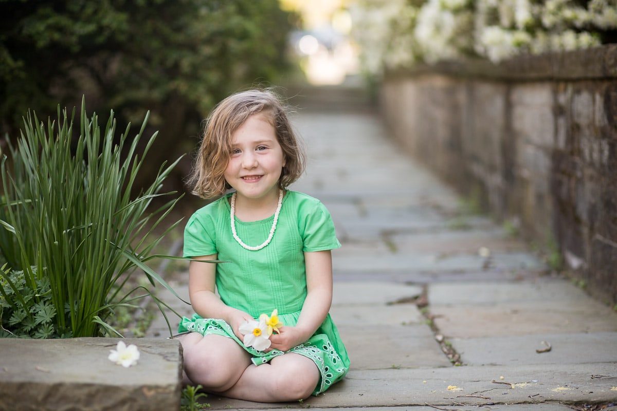 Young girl in green dress holding a flower sits on a stone walkway for solo and sibling photos on the grounds of Washington National Cathedral
