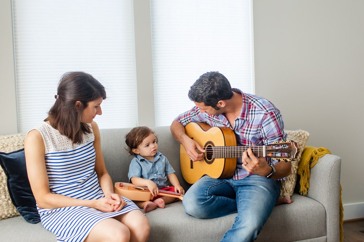 A mother and father sit together with baby son on couch while father plays guitar. When you choose a family photographer, discuss options for the location of the photo session, such as in the family's home where the baby can be in a comfortable, familiar environment.