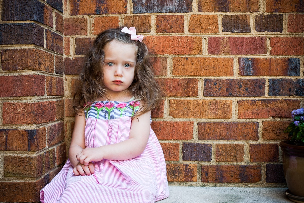 Image of little girl sitting in front of brick was was selected to display as a framed print in family's home. When choosing a family photographer, make sure they offer the end product you are looking for.