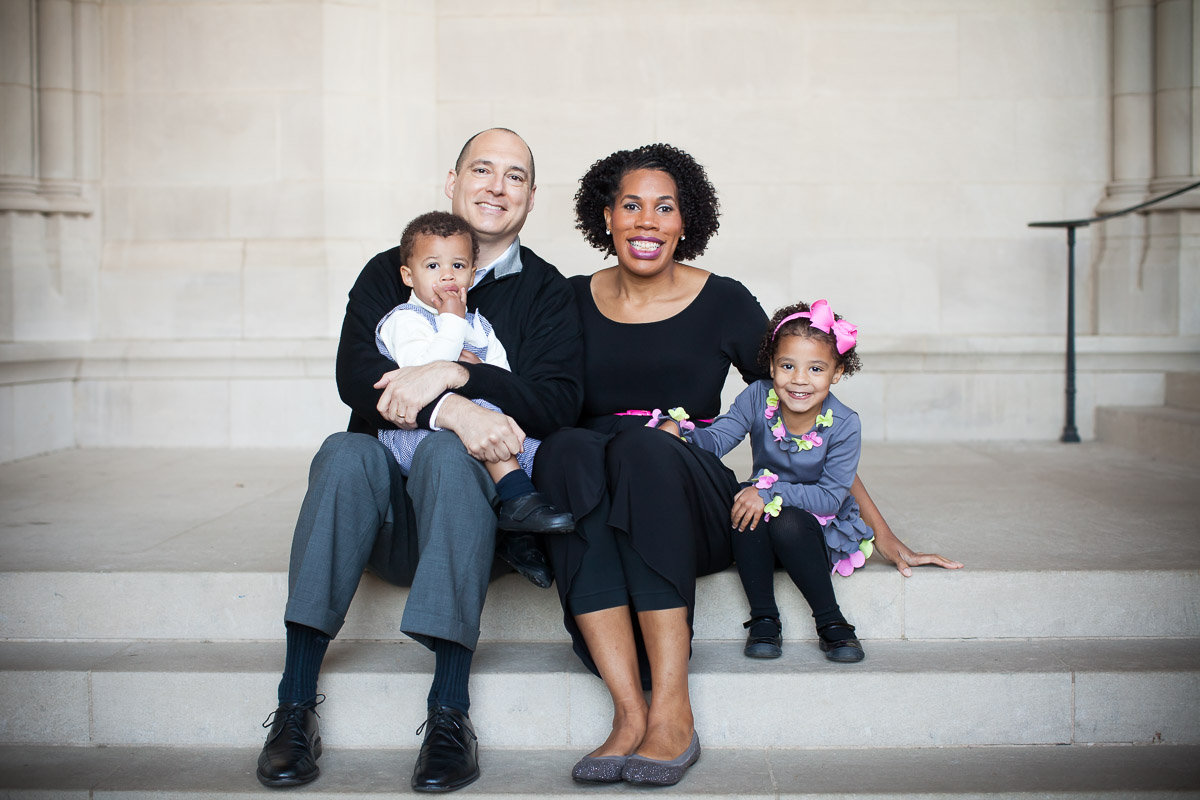 Family of four with parents, baby boy, and little girl sit together for a family photo on the steps outside Washington National Cathedral.